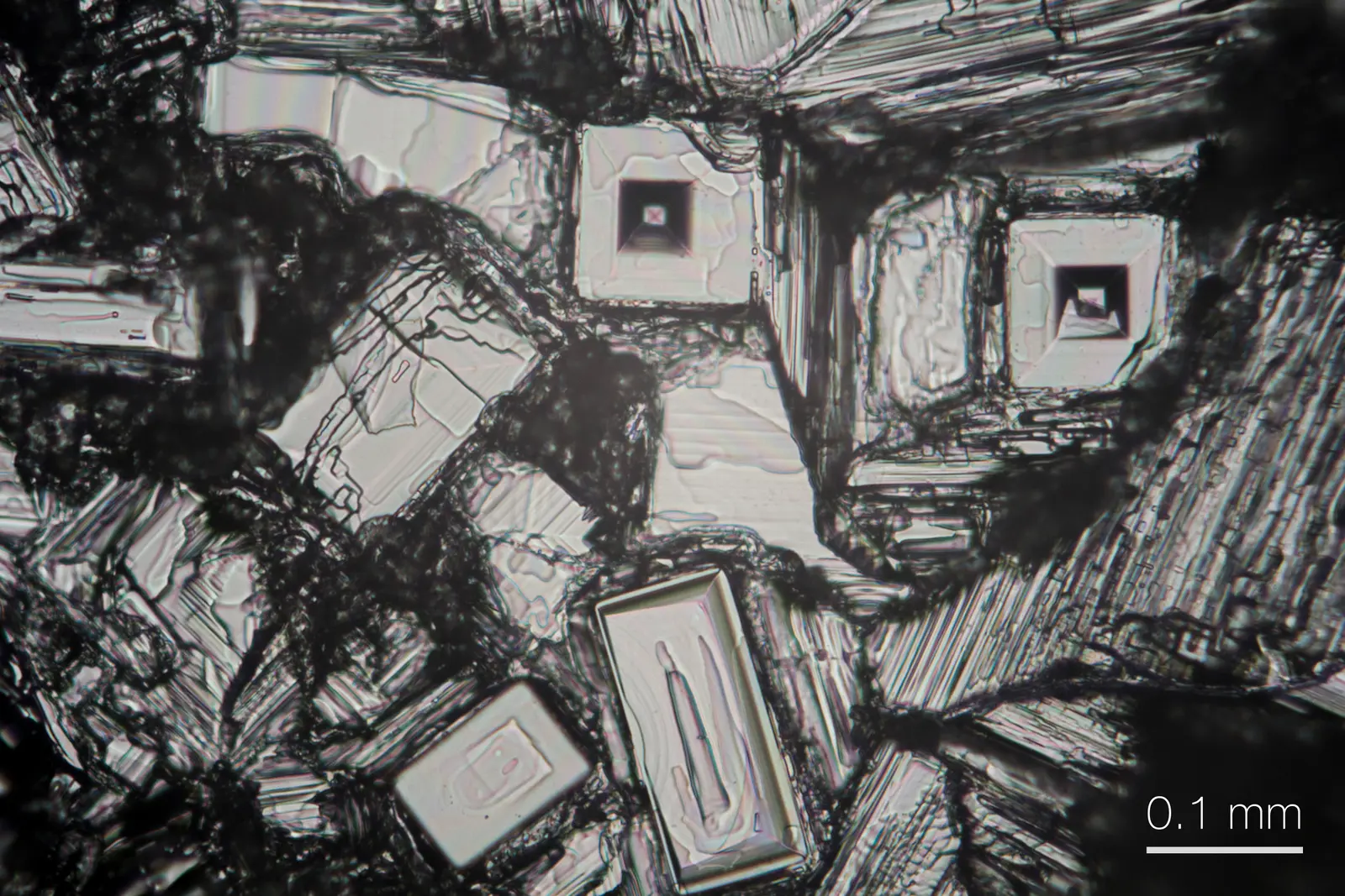 Sodium chloride crystals 0.08–0.19 mm across, light micrograph, native color; two square crystals with stepped, sunken centres; 0.1 mm bar.